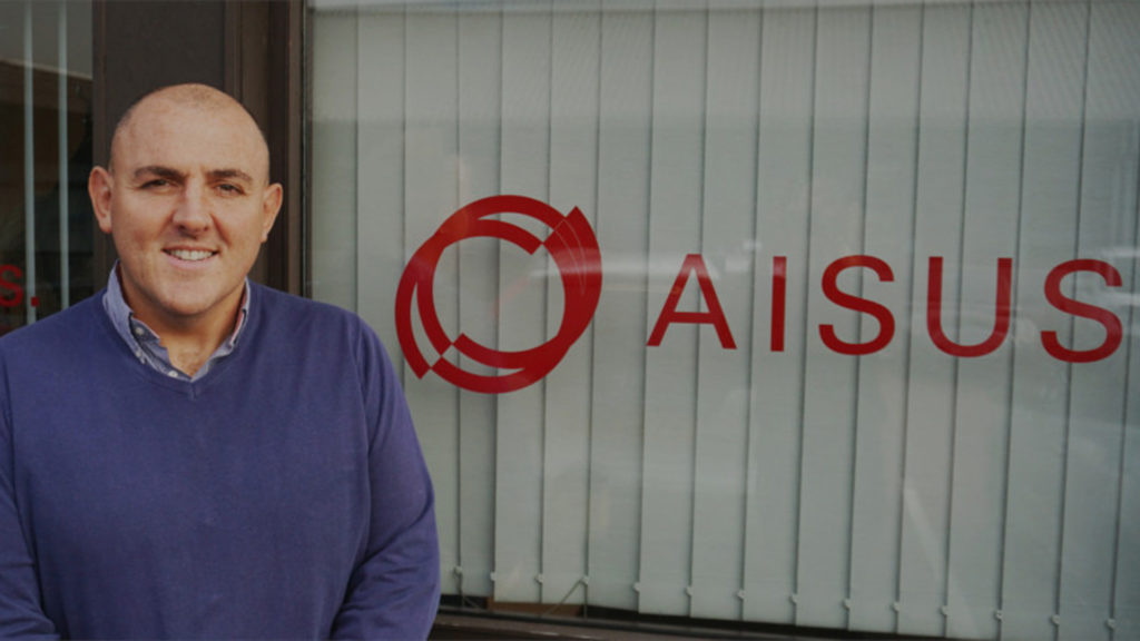smiling bald young man beside a red AISUS logo on a window